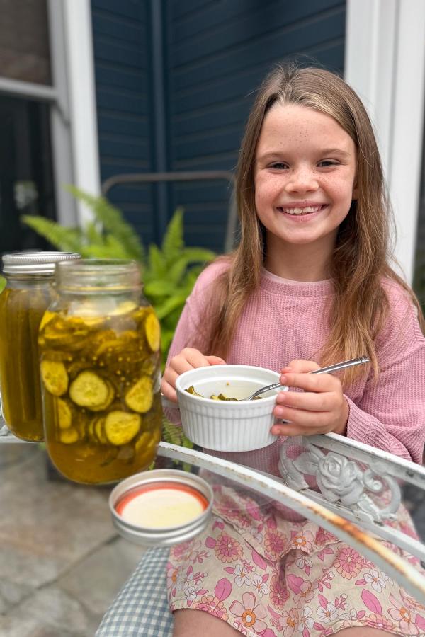 A smiling young girl in a pink shirt sits at a glass table outdoors, holding a bowl of pickles with a spoon. Two jars of sliced pickles are on the table beside her, and a blue house with plants is in the background.