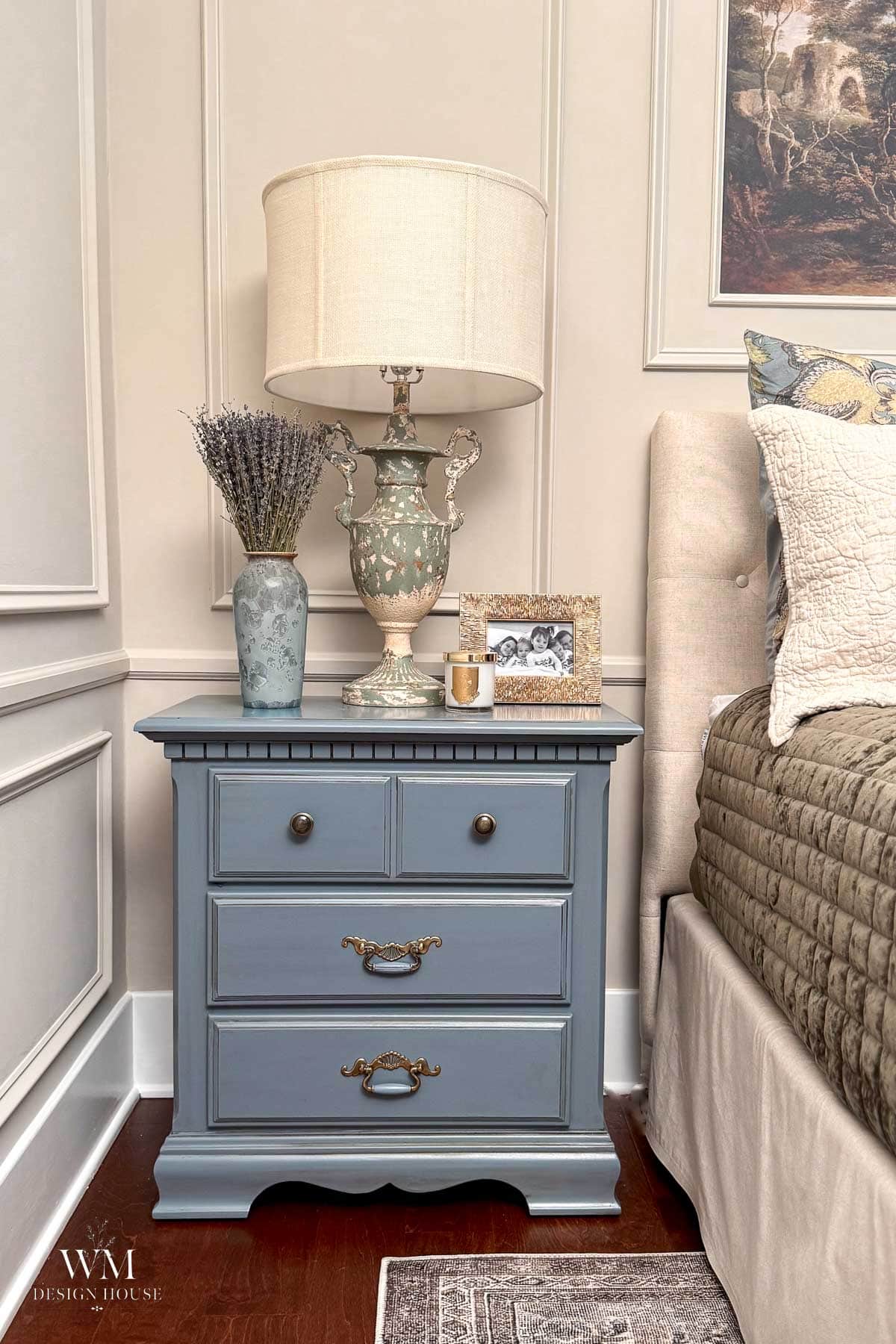 Blue nightstand with three drawers beside a bed, topped with a french lamp, a vase of lavender, a framed photo, and a candle. The room features classic decor and paneled walls painted in Benjamin Moore Smokey Taupe Paint.