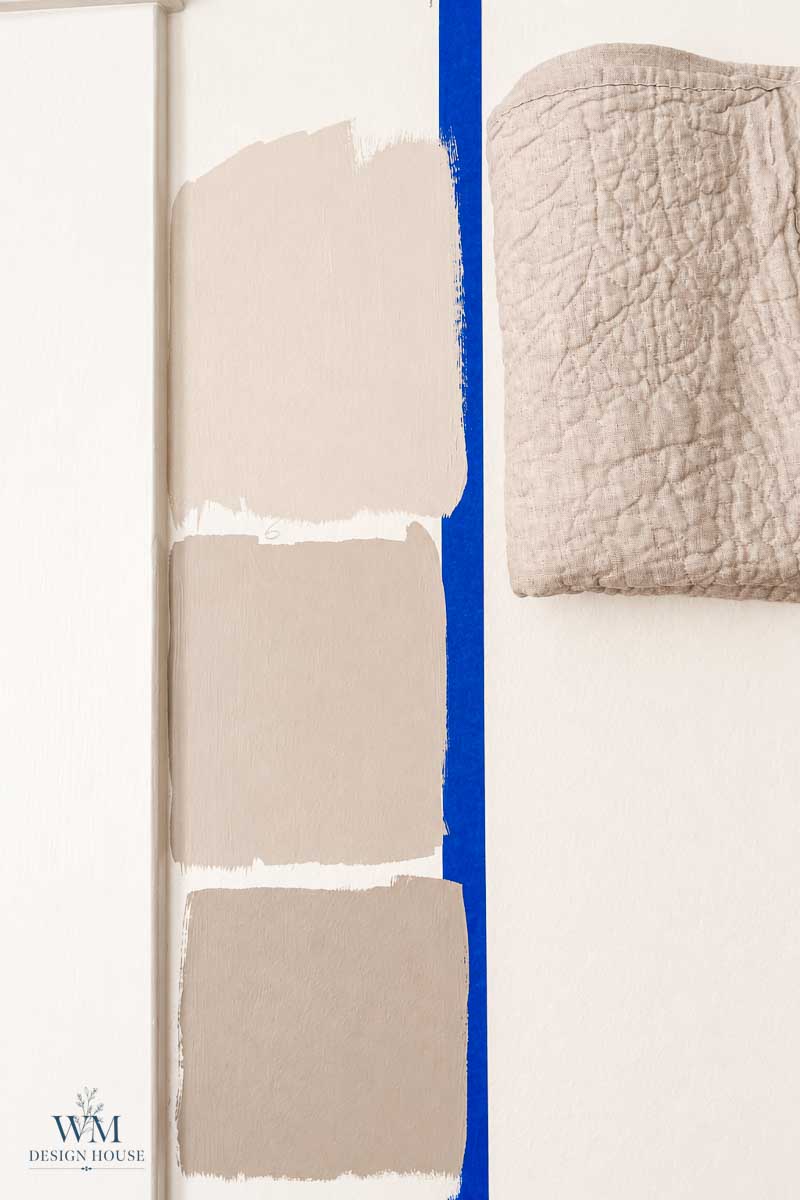 Three beige paint swatches, including Benjamin Moore Smokey Taupe Paint, are painted on a white wall, separated from a hanging beige quilt by a vertical strip of blue painter’s tape. The WM Design House logo is in the lower left corner.