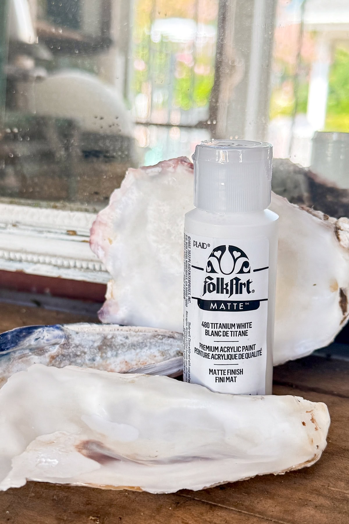 A bottle of FolkArt matte titanium white acrylic paint stands on a wooden surface in front of an open oyster shell, with a mirror and blurred outdoor scene in the background.
