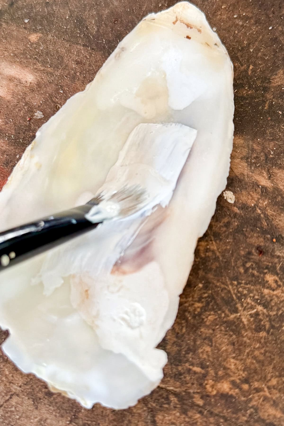 A paintbrush applies white paint onto a large, open oyster shell resting on a brown wooden surface.