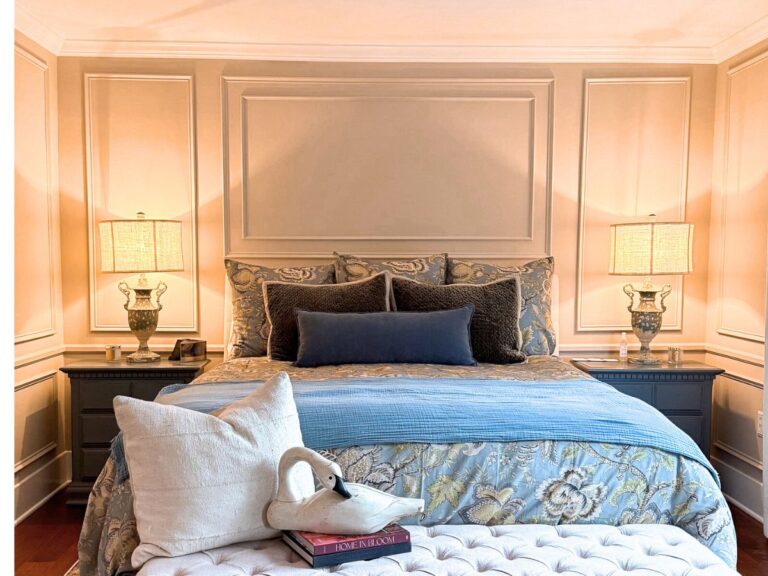 A neatly made bed with patterned bedding, a blue throw, and multiple pillows stands between two nightstands with matching lamps. DIY wall moldings add elegance above the headboard, while a bench holds a white pillow, swan ornament, and books.