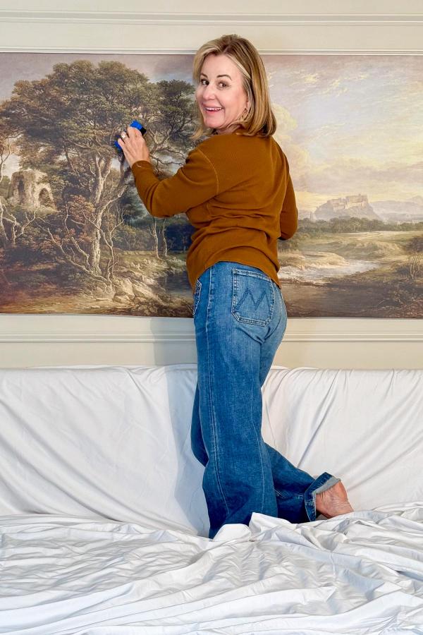 A woman stands barefoot on a bed, smiling at the camera while showing how to hang a wallpaper mural in the bedroom. She wears a brown sweater and blue jeans.