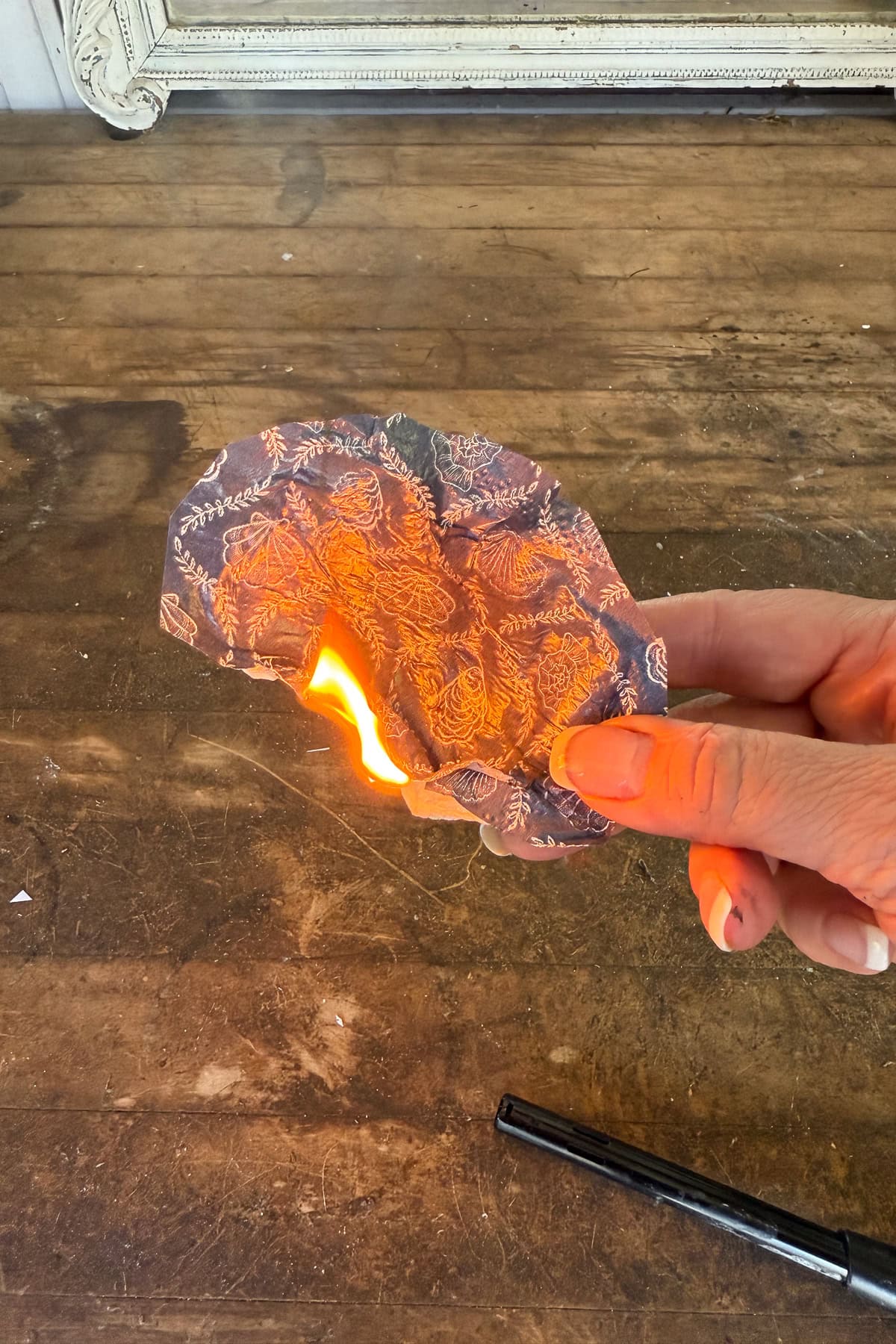 A hand holds a piece of floral-patterned fabric that is burning at one edge, with a lighter resting on a wooden surface below.