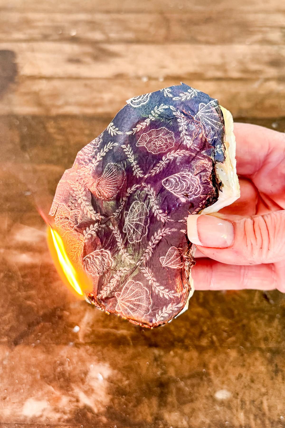 A hand holds a piece of wood with a decorative pattern of leaves and pinecones burning on one edge, with flames and scorch marks visible against a wooden surface background.