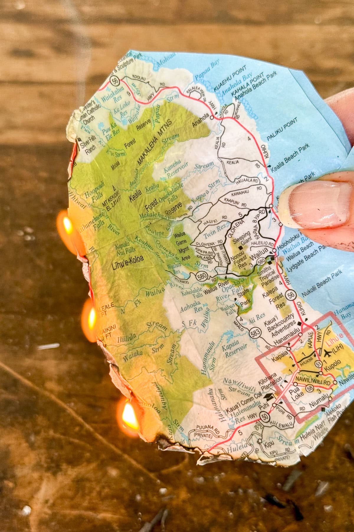 A person holds a torn piece of a colorful map with flames burning its edge, set against a wooden background.