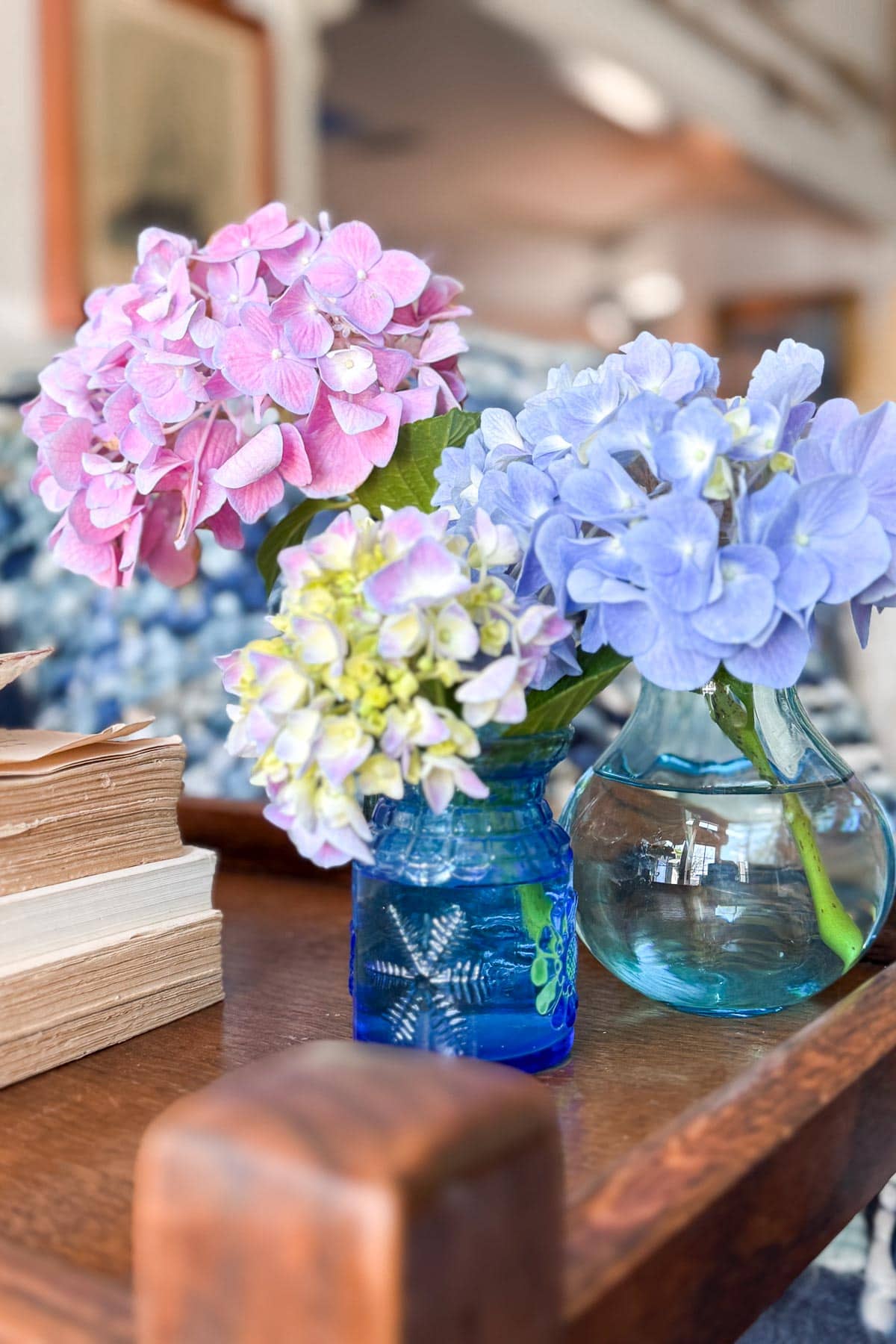 Coastal Cottage Table Decor-Two glass vases with pink and blue hydrangea flowers are placed on a wooden surface next to a stack of books, with a softly blurred background of a cozy indoor setting.