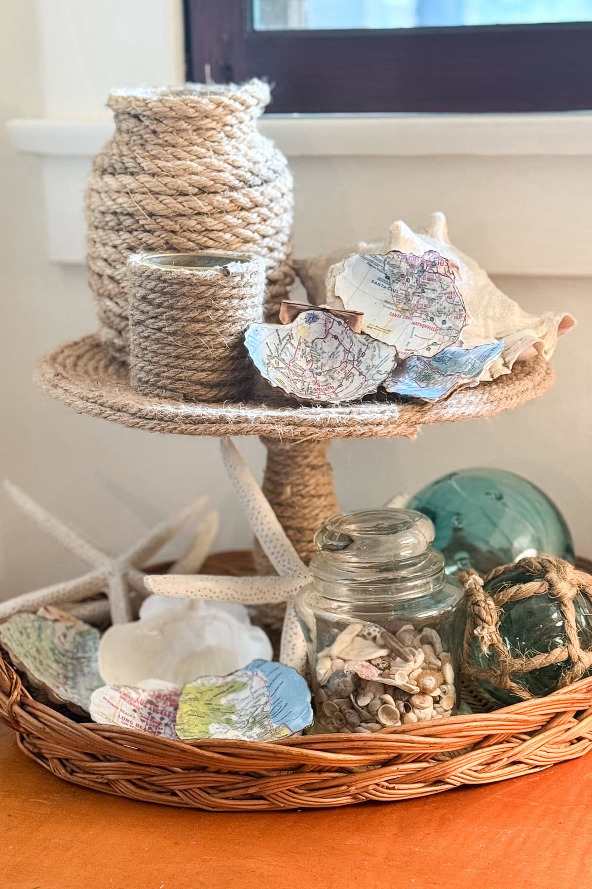 A two-tiered wicker stand displays rope-wrapped jars, seashells, starfish, and glass jars filled with shells. Pieces of paper with map prints are scattered among the beach-themed decor.