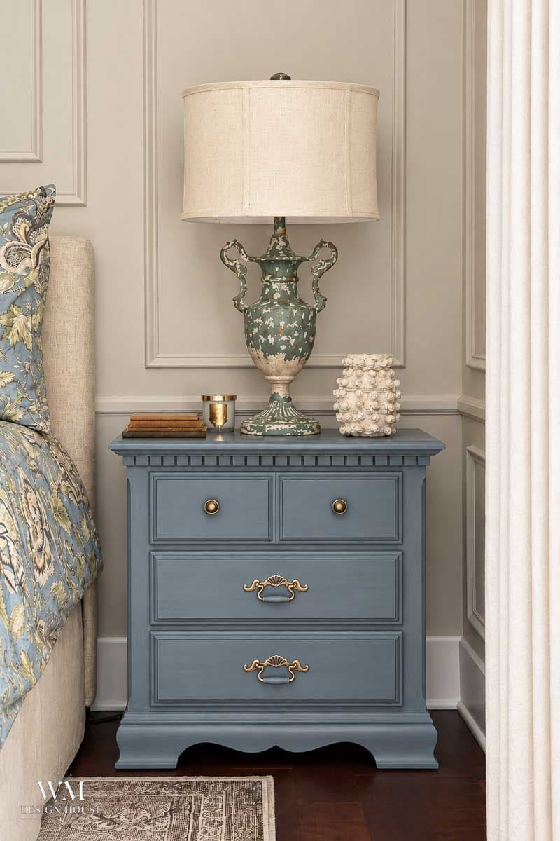Blue bedside table with three drawers, a decorative green and cream lamp, a white textured ornament, a gold cup, and a book on top—set beside a beige bed and against a cream paneled wall painted in Benjamin Moore Smokey Taupe Paint.