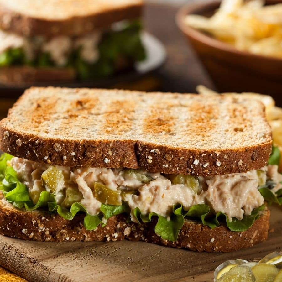 A tuna salad sandwich with lettuce and Mrs. Wages Bread and Butter Pickles on multigrain bread sits on a wooden board, with another sandwich and a bowl of potato chips blurred in the background.