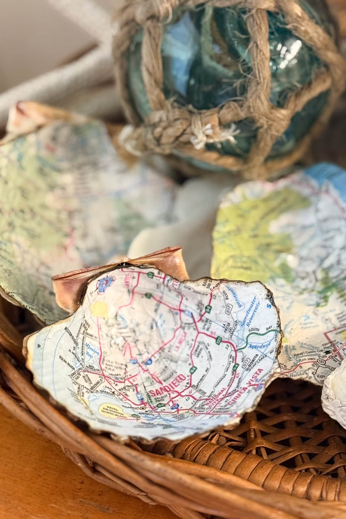 A wicker basket contains decorative clam shells decoupaged with map prints. Behind them is a glass fishing float wrapped in rope. The scene has a coastal, nautical theme.