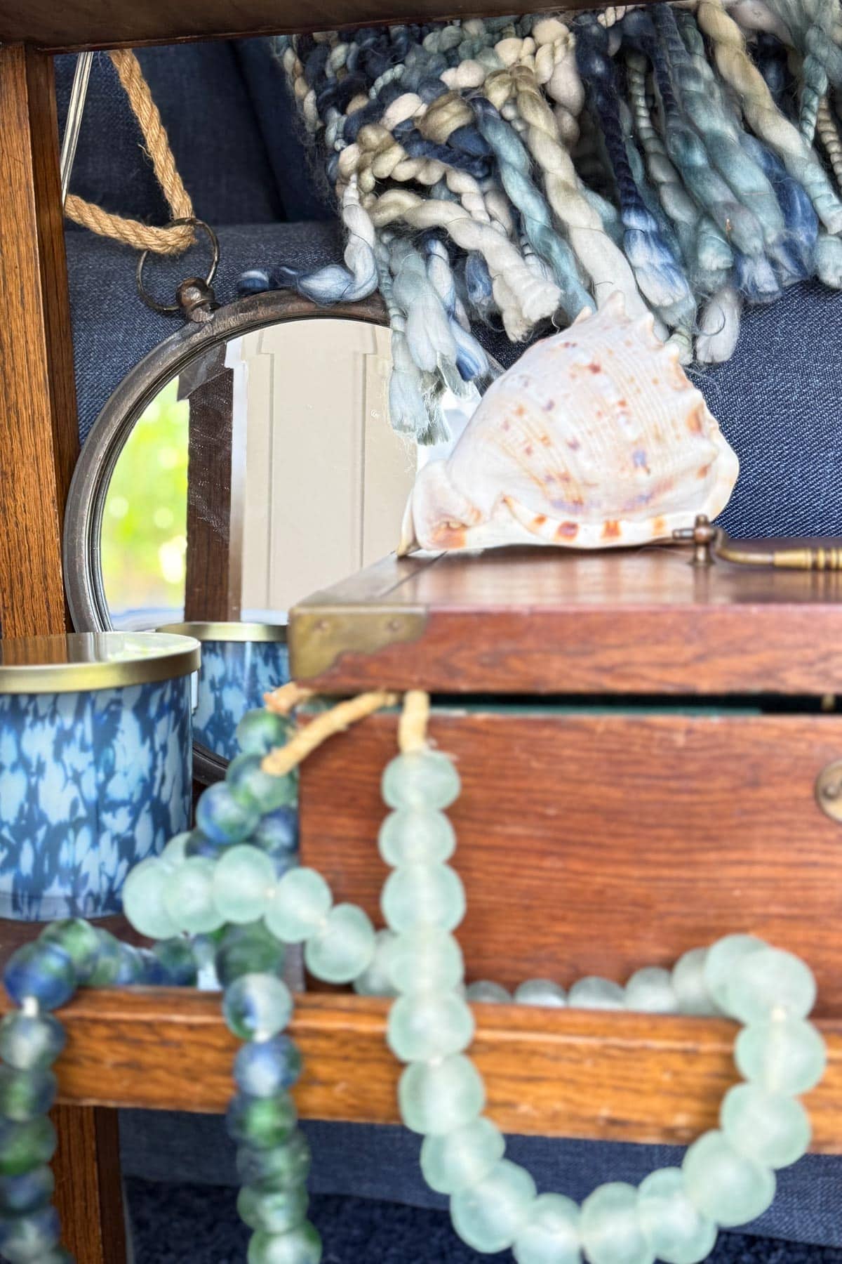 A wooden shelf displays blue and green beaded necklaces, a wooden box, a seashell, a small round mirror, and blue-patterned containers. A folded, textured fabric lies in the background.