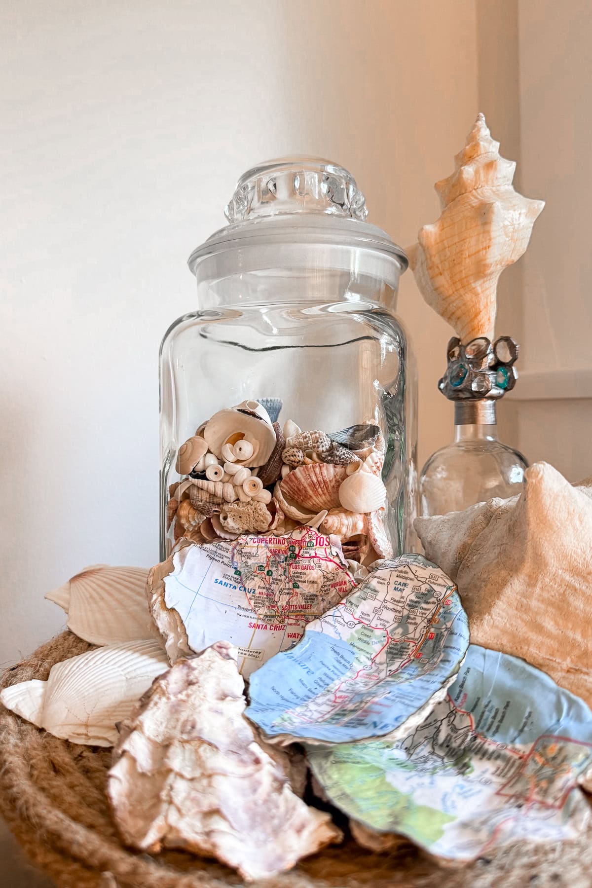 A glass jar filled with seashells sits on a woven mat, surrounded by larger seashells and oyster shells painted with colorful maps. A decorative conch shell and bottle are in the background.