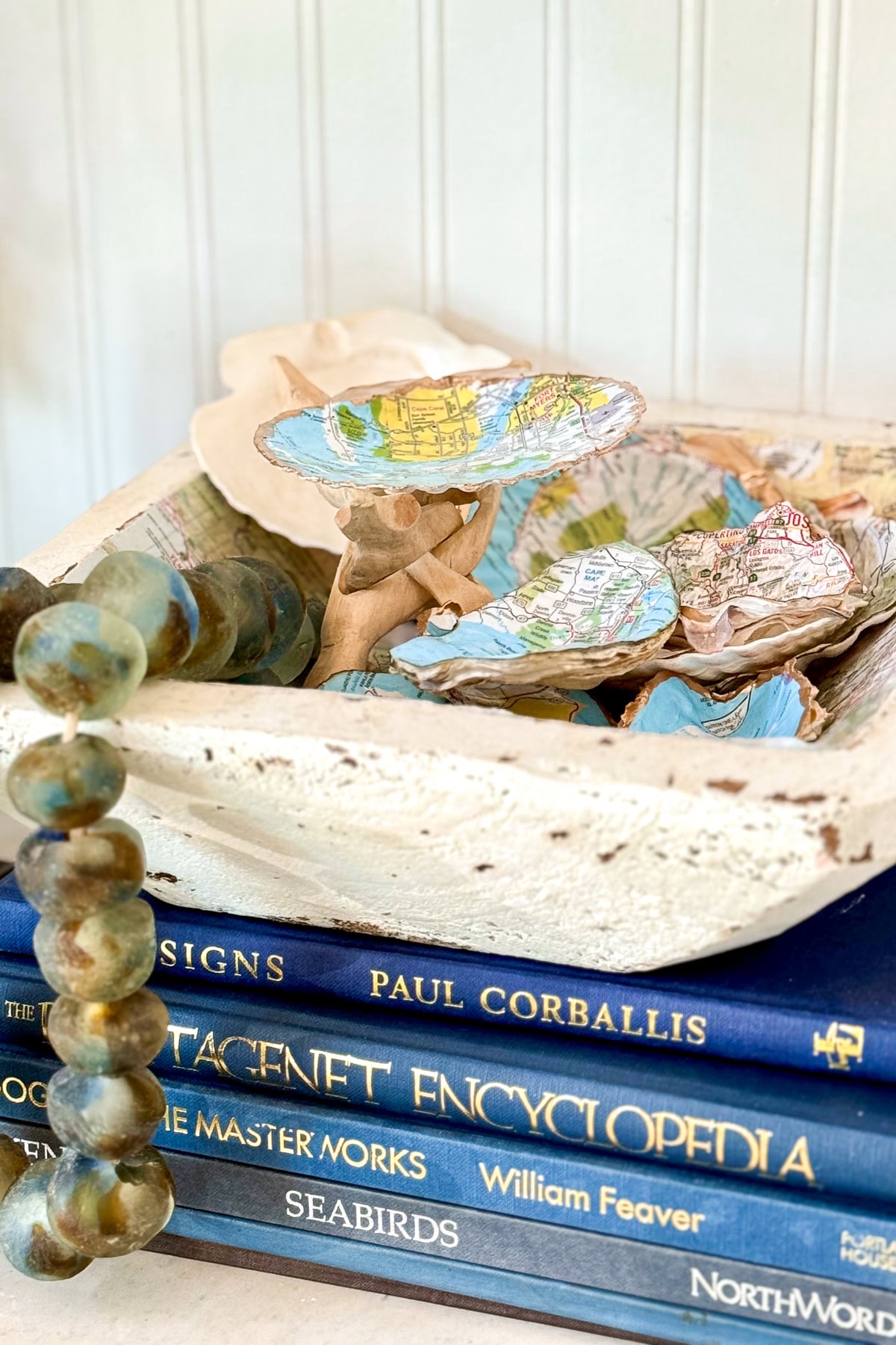 A decorative bowl filled with seashells lined with map fragments sits on top of a stack of hardcover books, including titles about signs, encyclopedias, master works, and seabirds. Draped beads are also visible.