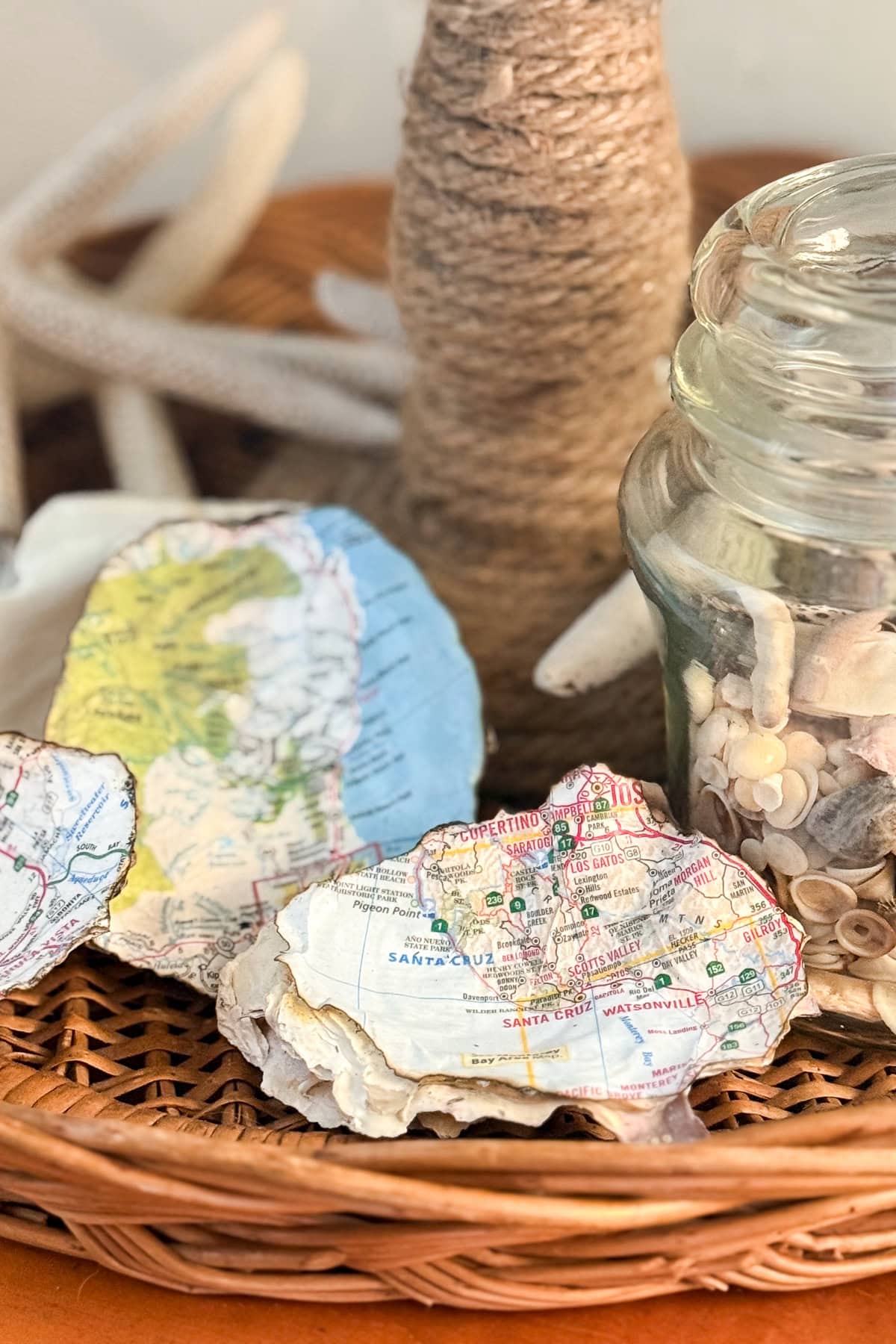 A close-up of a wicker tray holding map-decorated seashells, a glass jar filled with seashells, and a twine-wrapped bottle. In the background, white coral is partially visible.