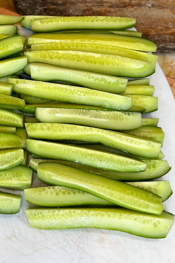 Several cucumber spears are neatly arranged in rows on a white cutting board, with their green skins and pale, seeded interiors visible.