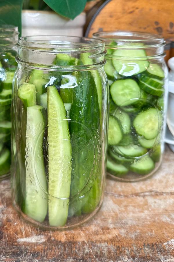Two glass jars on a wooden surface: one filled with cucumber spears and the other with sliced cucumber rounds, both in clear liquid.