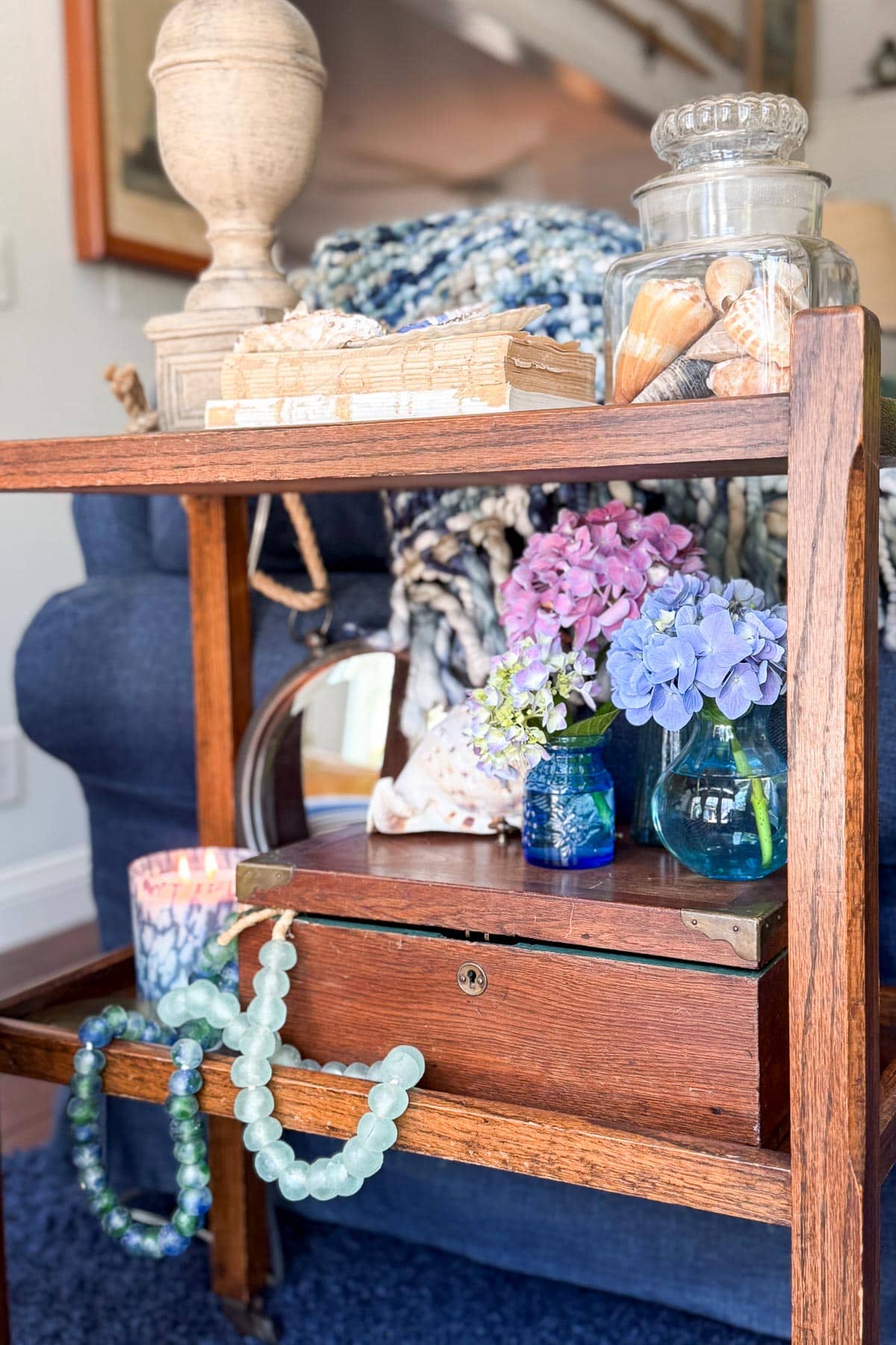Coastal Cottage Table Decor-A wooden side table displays glass bead necklaces, blue and pink hydrangeas in small vases, a candle, seashells in a glass jar, vintage books, and a textured blanket draped over a blue armchair in the background.