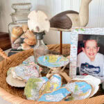 A wicker tray holds seashells lined with map paper, a wood bird figurine, glass jars with shells, a white sand dollar, and a framed photo of a smiling boy in front of a map background.