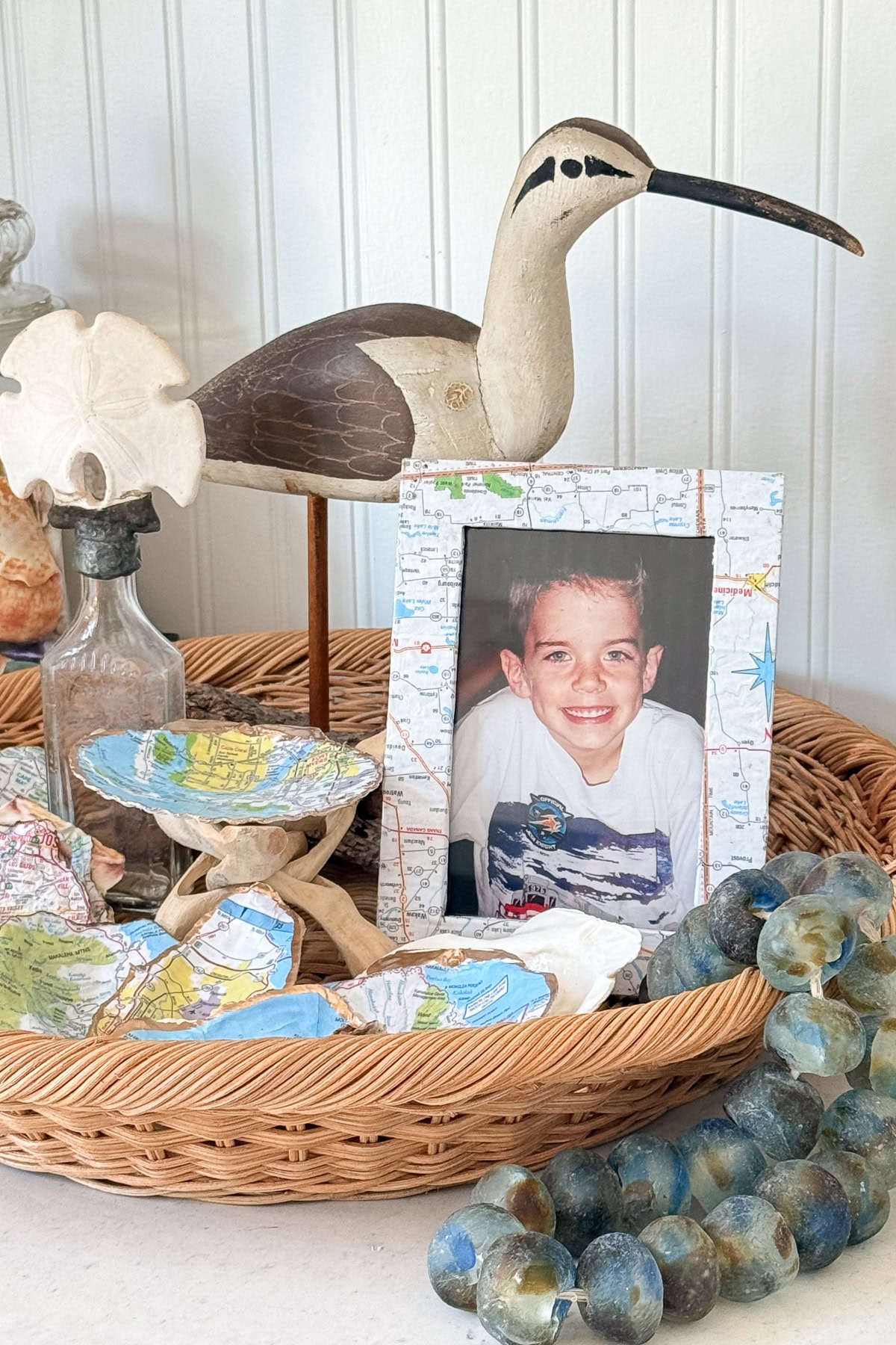 A wicker tray holds a framed photo of a smiling boy, a wooden bird sculpture, a bottle, decorative globes, and small map-covered bowls. The background is white beadboard paneling.