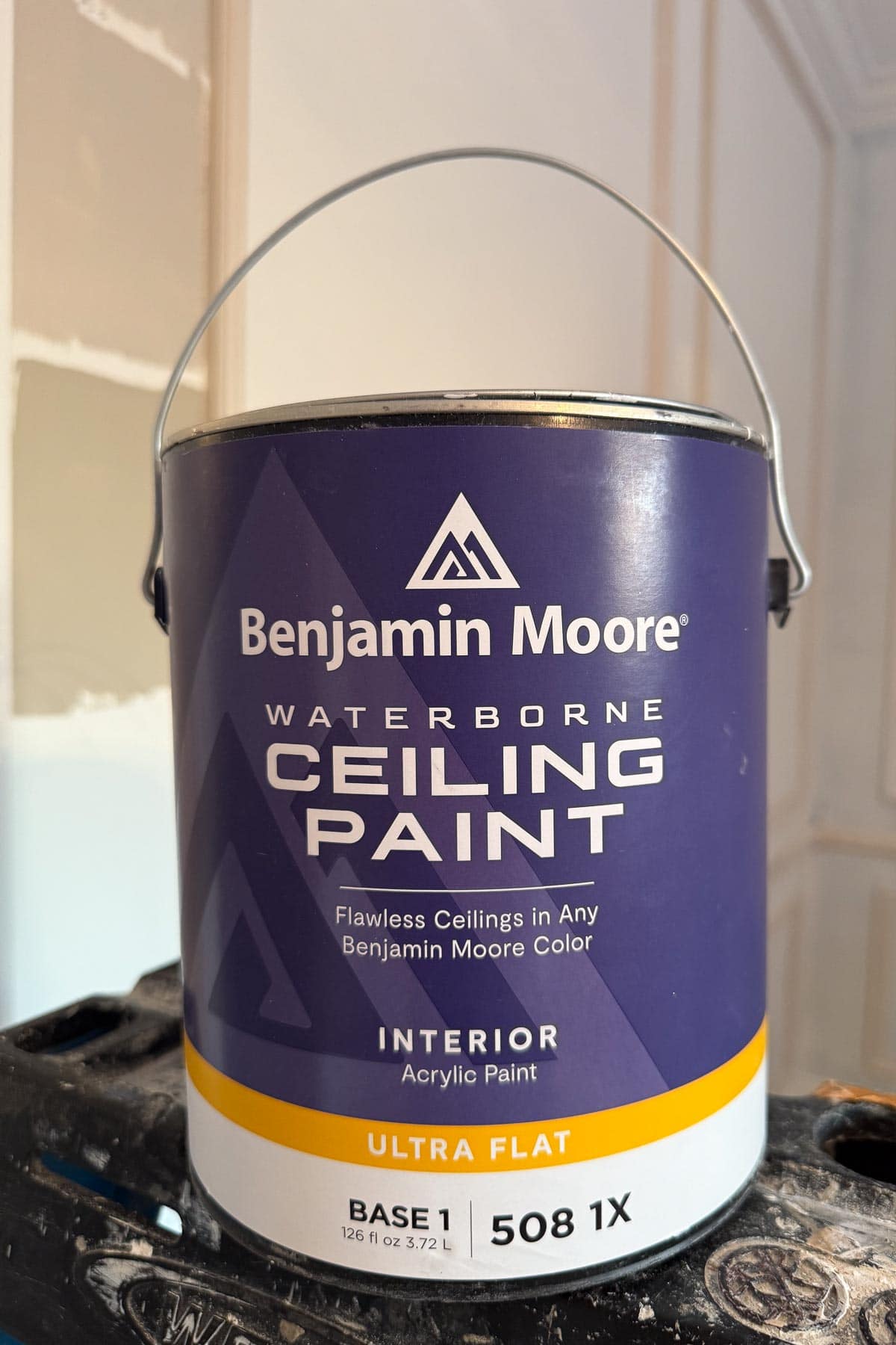 A can of Benjamin Moore Waterborne Ceiling Paint, labeled as interior, ultra flat, base 1, 508 1X, sitting on a black surface with a partially painted wall in the background.