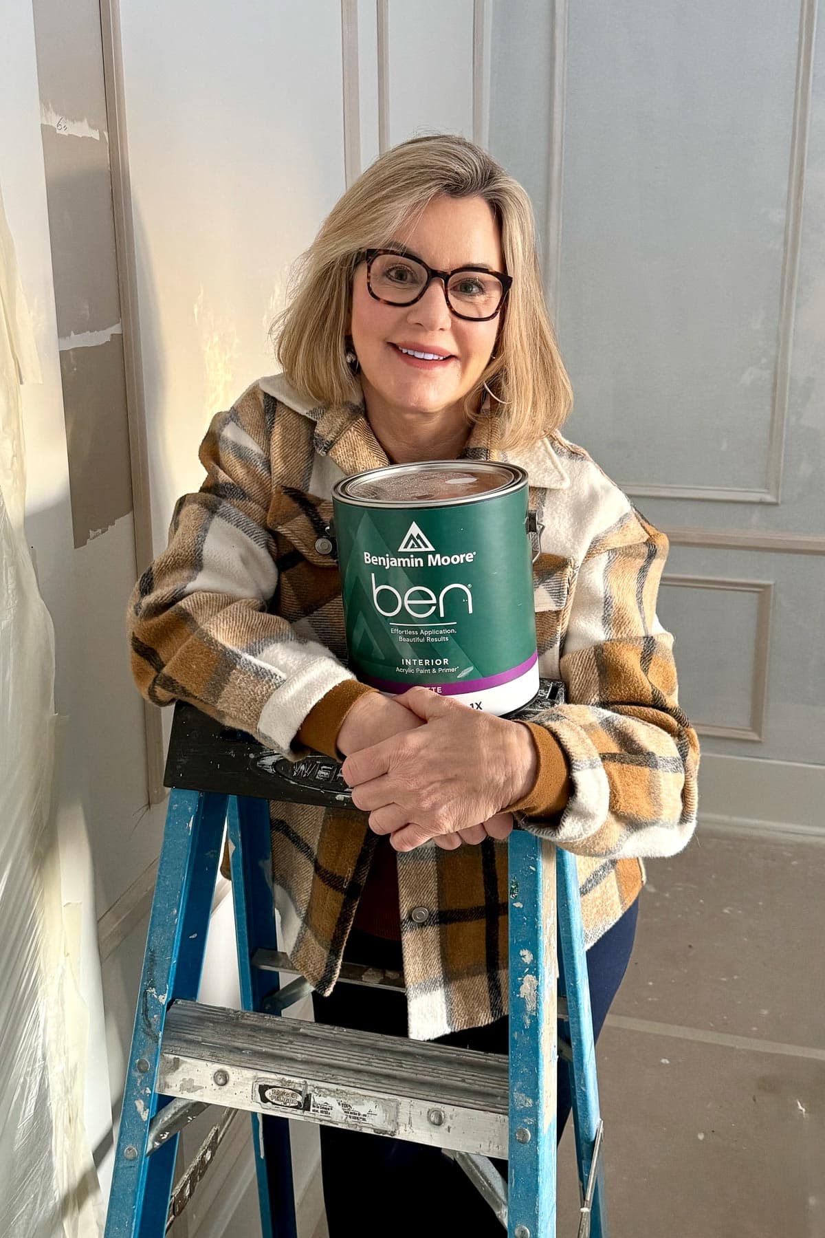 A woman in glasses and a plaid jacket smiles while leaning on a blue ladder, holding a can of Benjamin Moore paint in a room with light-colored walls.