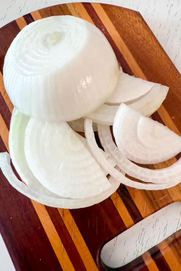 A partially sliced white onion sits on a wooden cutting board with several onion rings and slices arranged beside it.