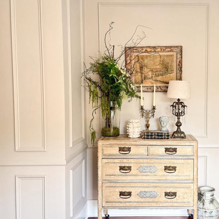 An ornate dresser with decorative handles holds a tall vase of green branches, candles, books, and small decor. Above it hangs framed artwork on a light-colored wall enhanced with elegant DIY Wall Moldings.