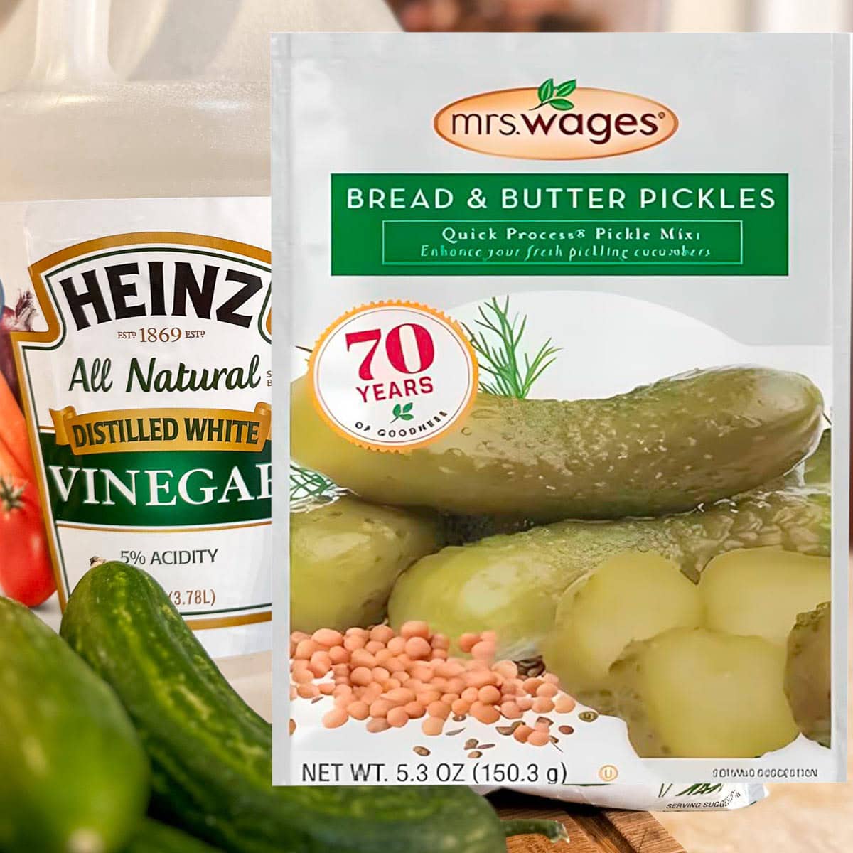 A packet of Mrs. Wages Bread and Butter Pickles Mix sits in front of a Heinz distilled white vinegar bottle, fresh cucumbers, and mustard seeds. The pickle mix packet highlights "70 years of goodness.