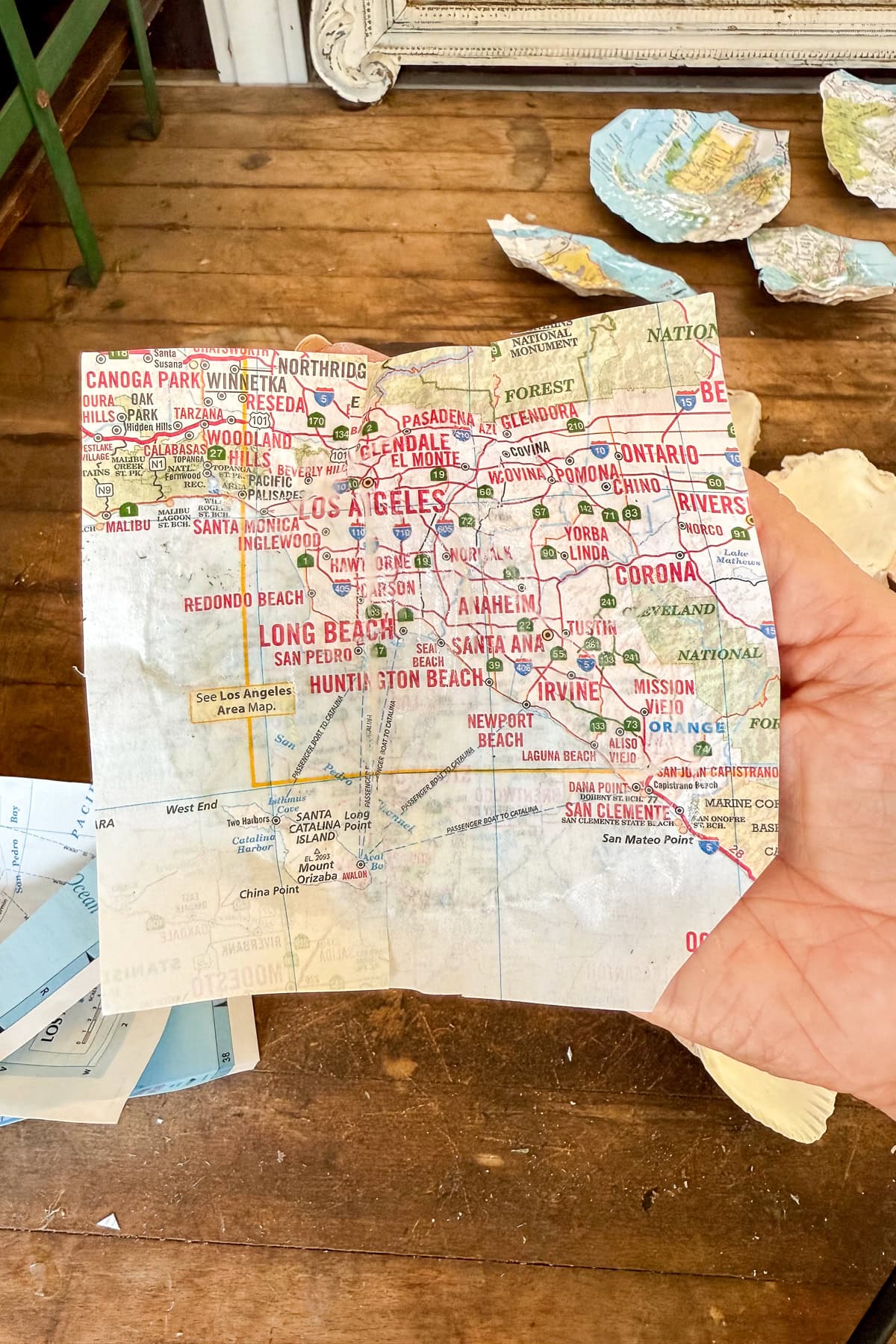 A hand holds a crumpled map focusing on Los Angeles and surrounding cities in Southern California, with several other torn map pieces scattered on a wooden floor in the background.