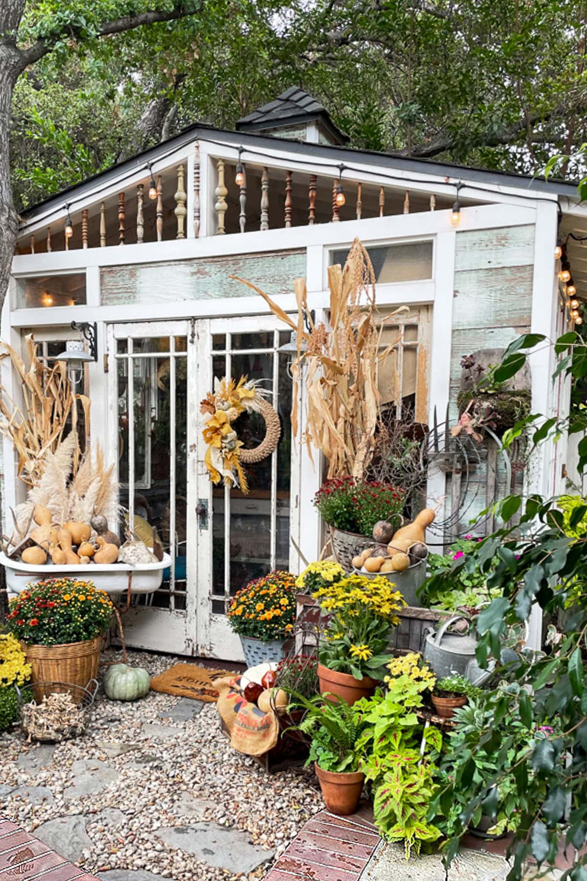 A rustic garden shed decorated for autumn, inspired by The Fall Shop, with dried cornstalks, pumpkins, gourds, potted plants, flowers, and a wreath on the door. Baskets and pots filled with fall harvest items surround the entrance.