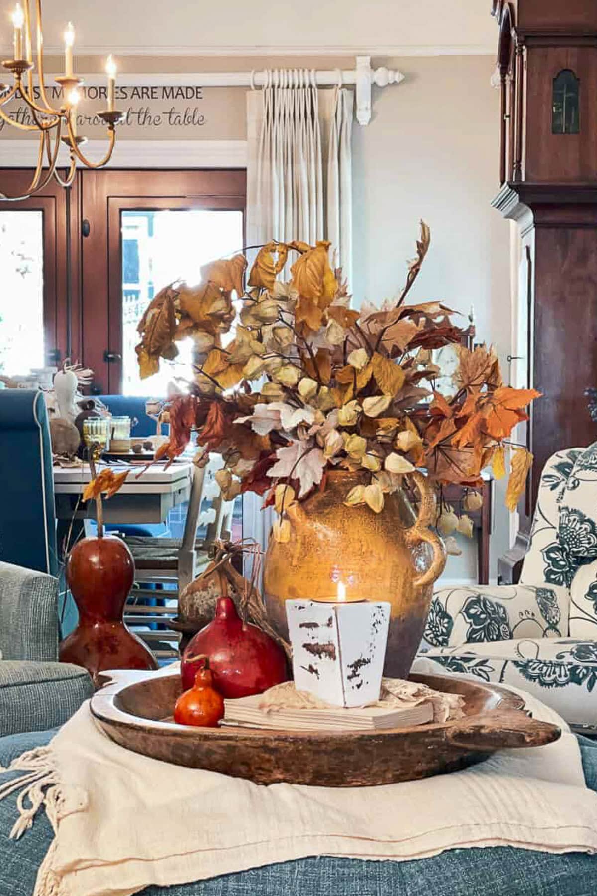 A cozy living room scene features a wooden tray with a candle, two gourds, and a pomegranate on a coffee table. A large vase with dried autumn leaves from The Fall Shop serves as the centerpiece, surrounded by neutral furnishings.