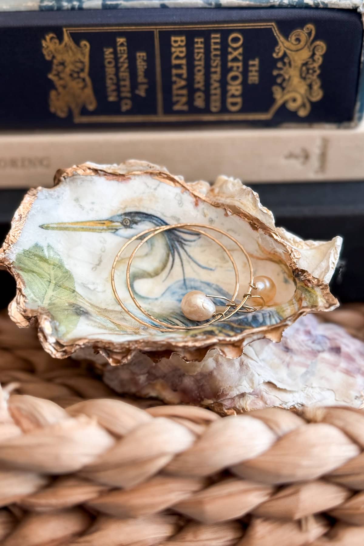 A painted oyster shell featuring a heron holds gold hoop earrings with pearls. The shell rests on a woven surface, with books, including "The Oxford Illustrated History of Britain," in the background.