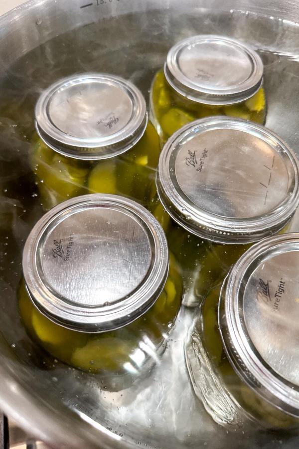 Jars filled with green vegetables are being processed in hot water inside a large pot, likely for canning or preserving. The metal lids are secured as the jars sit partially submerged in the water.
