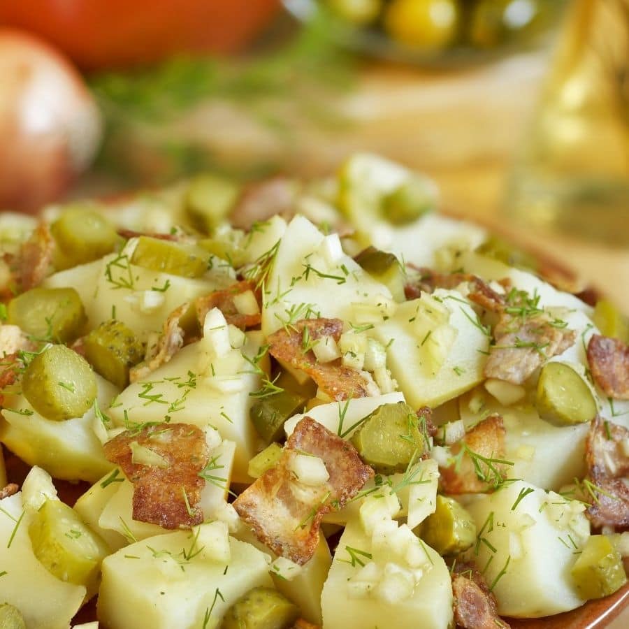 A close-up of a potato salad topped with Mrs. Wages Bread and Butter Pickles, crispy bacon pieces, and fresh dill, with blurred vegetables in the background.
