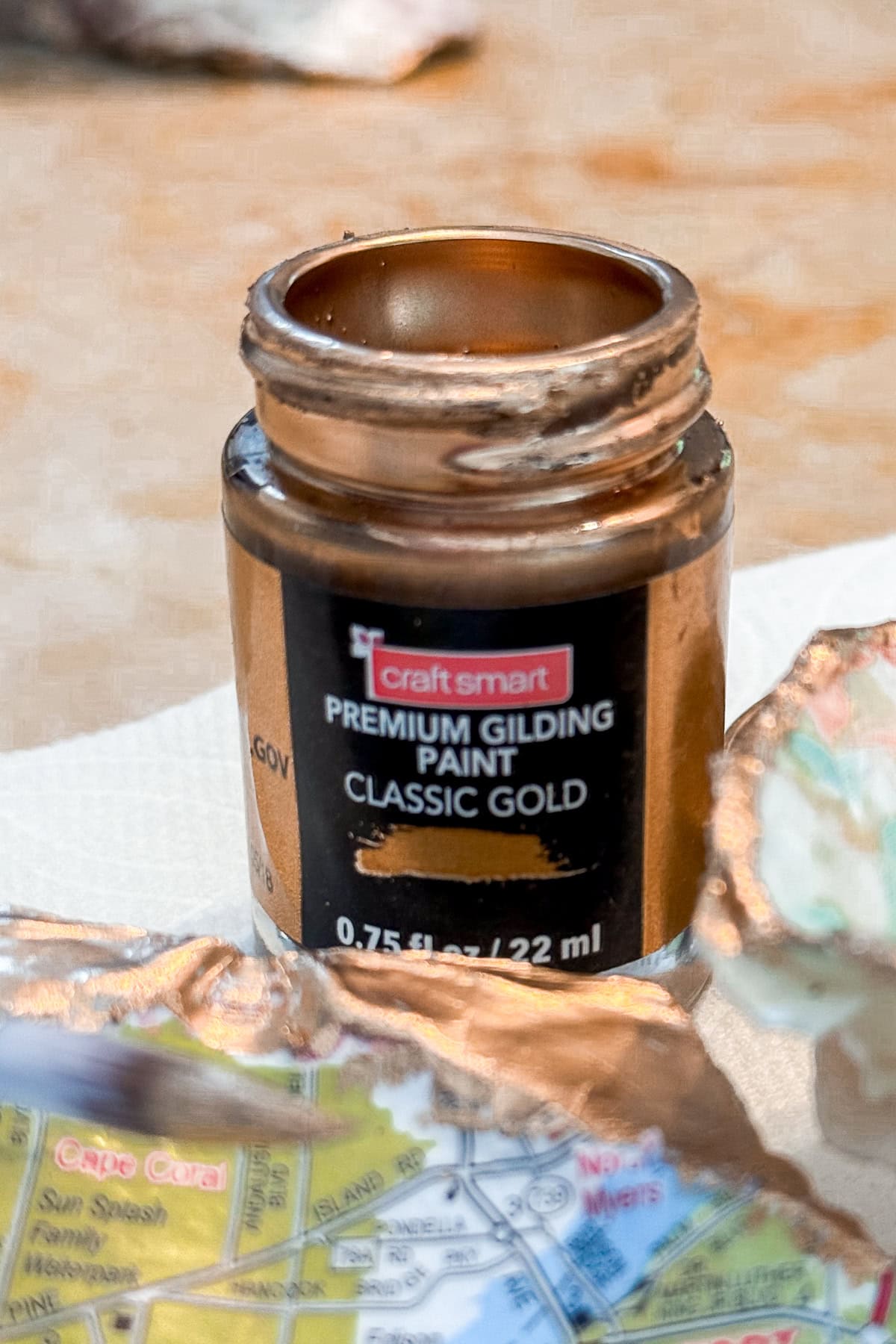 An open jar of Craft Smart Premium Gilding Paint in Classic Gold sits on a surface, gold paint smeared around the rim, with Decoupage Shells with Maps and a partially painted paper map nearby.