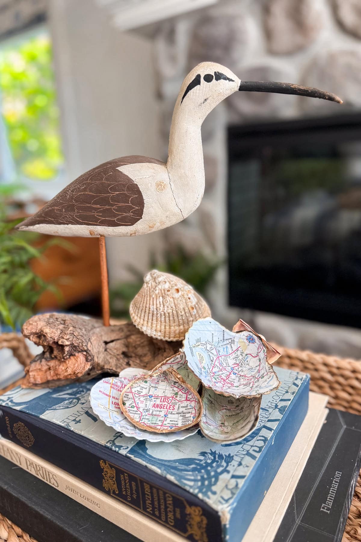 A wooden shorebird figurine stands on a table next to seashells, including some decoupaged with colorful map designs, arranged on top of a stack of books in a cozy, light-filled room.
