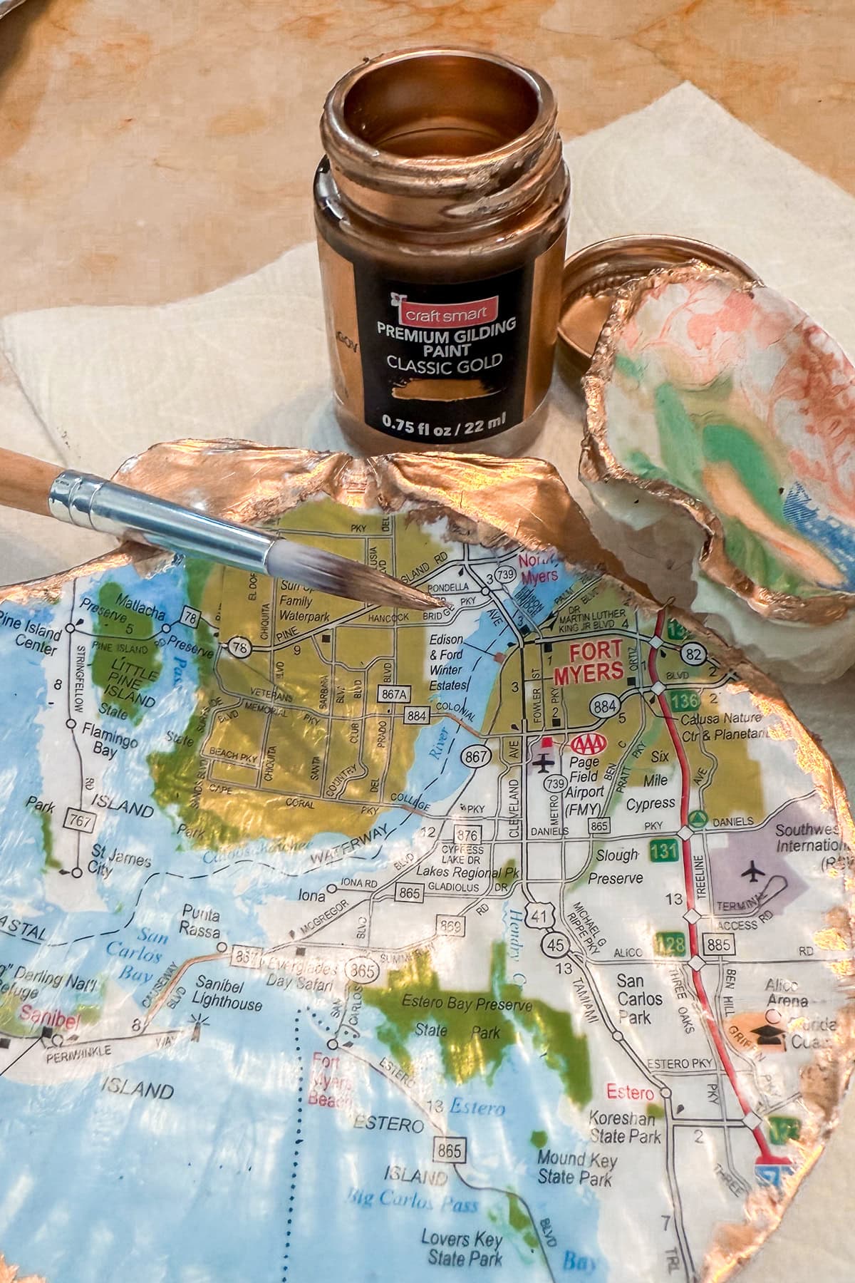 A decorative oyster shell, part of Decoupage Shells with Maps, is lined with a map of Fort Myers, Florida and trimmed in gold. It sits beside a gold gilding paint jar, paintbrush, and painted seashell on a tan surface.