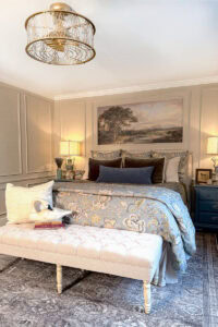 A cozy master bedroom features a tufted bench at the foot of a bed with patterned bedding and velvet pillows, flanked by blue nightstands, table lamps, and a large landscape painting—perfect inspiration for master bedroom decorating ideas.