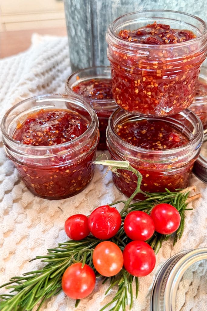 Four glass jars filled with homemade tomato jam are stacked on a textured cloth. In front of the jars, a sprig of rosemary holds several red and orange cherry tomatoes.