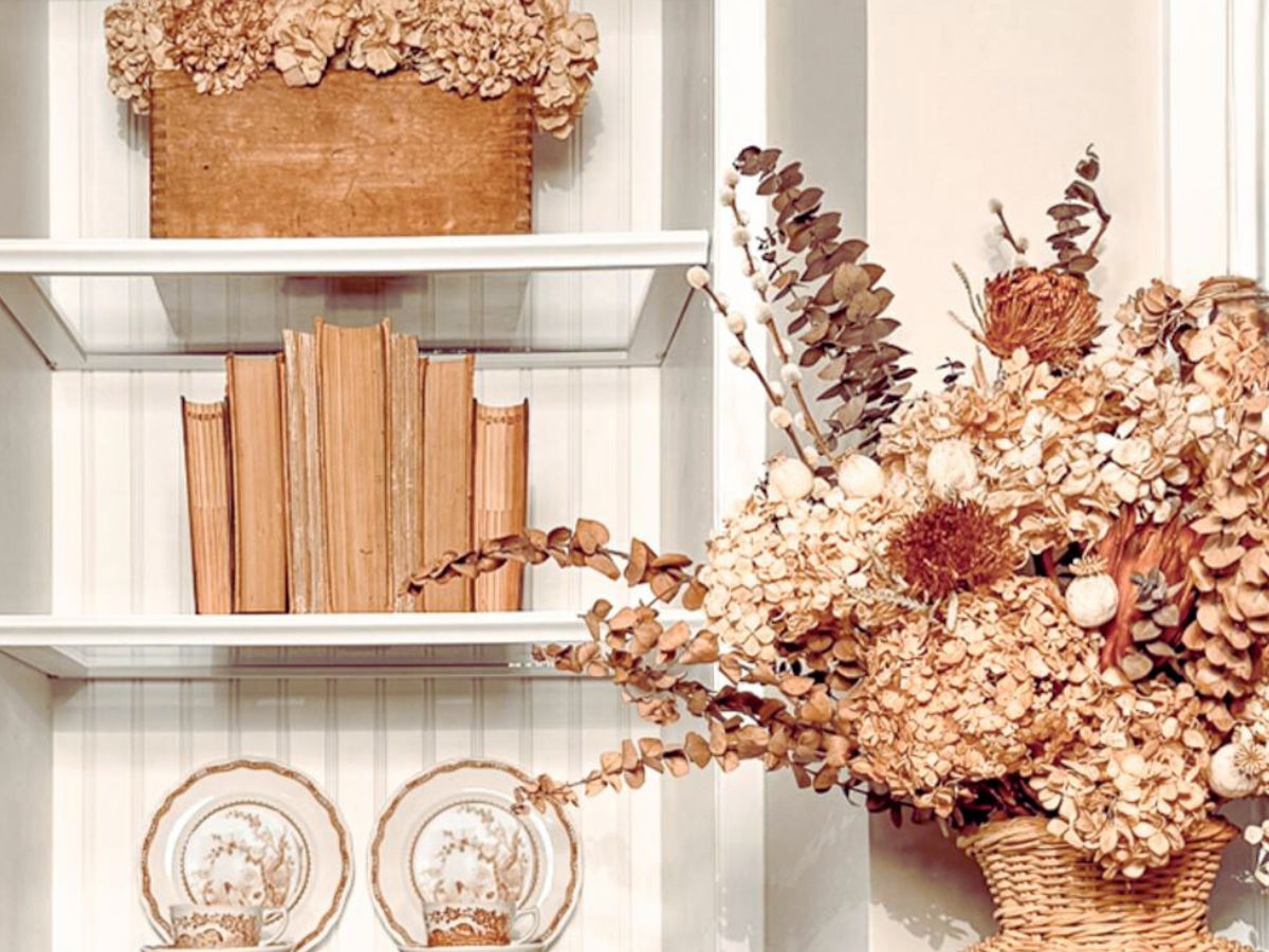 A white shelf displays a wooden box with dried flowers, vintage books, and decorative plates—a beautiful example of budget friendly fall shelf styling with thrifted decor in neutral, earthy tones.