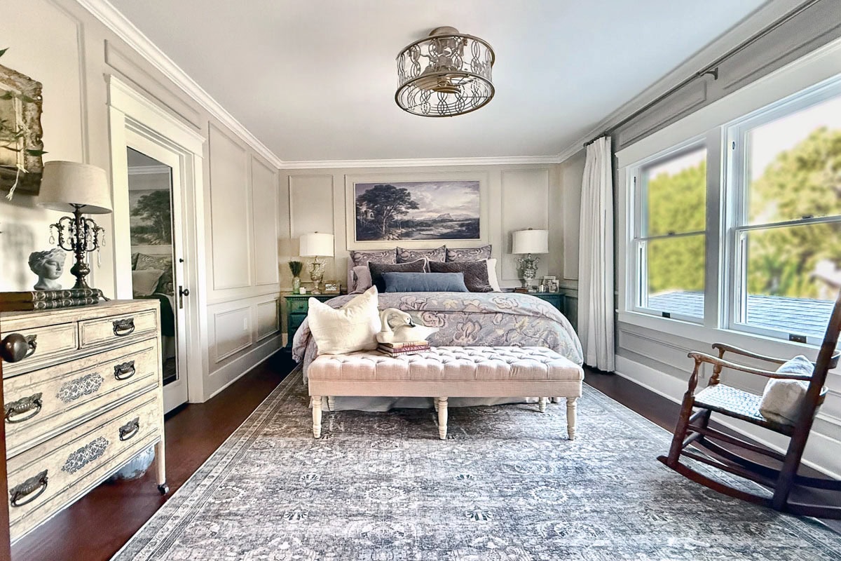 Elegant bedroom with neutral tones, featuring a large bed with decorative pillows, a tufted bench at the foot, and a patterned rug—perfect inspiration for those seeking master bedroom decorating ideas. Three large windows fill the space with natural light.