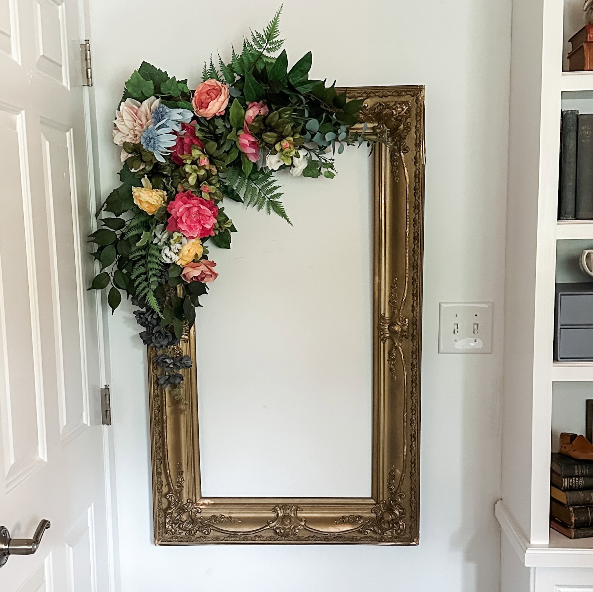 A gold ornate mirror frame hangs on a wall. Artificial flowers and greenery are arranged on the top left corner, cascading down the frame. The mirror has no glass, with wall and shelves visible through it.