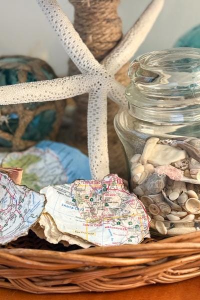 A wicker basket holds seashells, a glass jar of shells, a white starfish, and torn map pieces, creating a coastal-themed decorative arrangement.