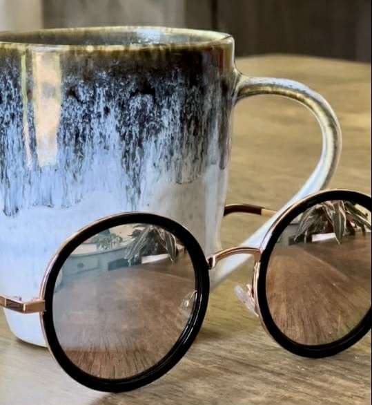 A ceramic mug with a textured, dark-to-light glaze sits on a wooden surface behind round eyeglasses, which reflect part of the room in their lenses. Steam rises from the mug, suggesting a hot beverage inside.