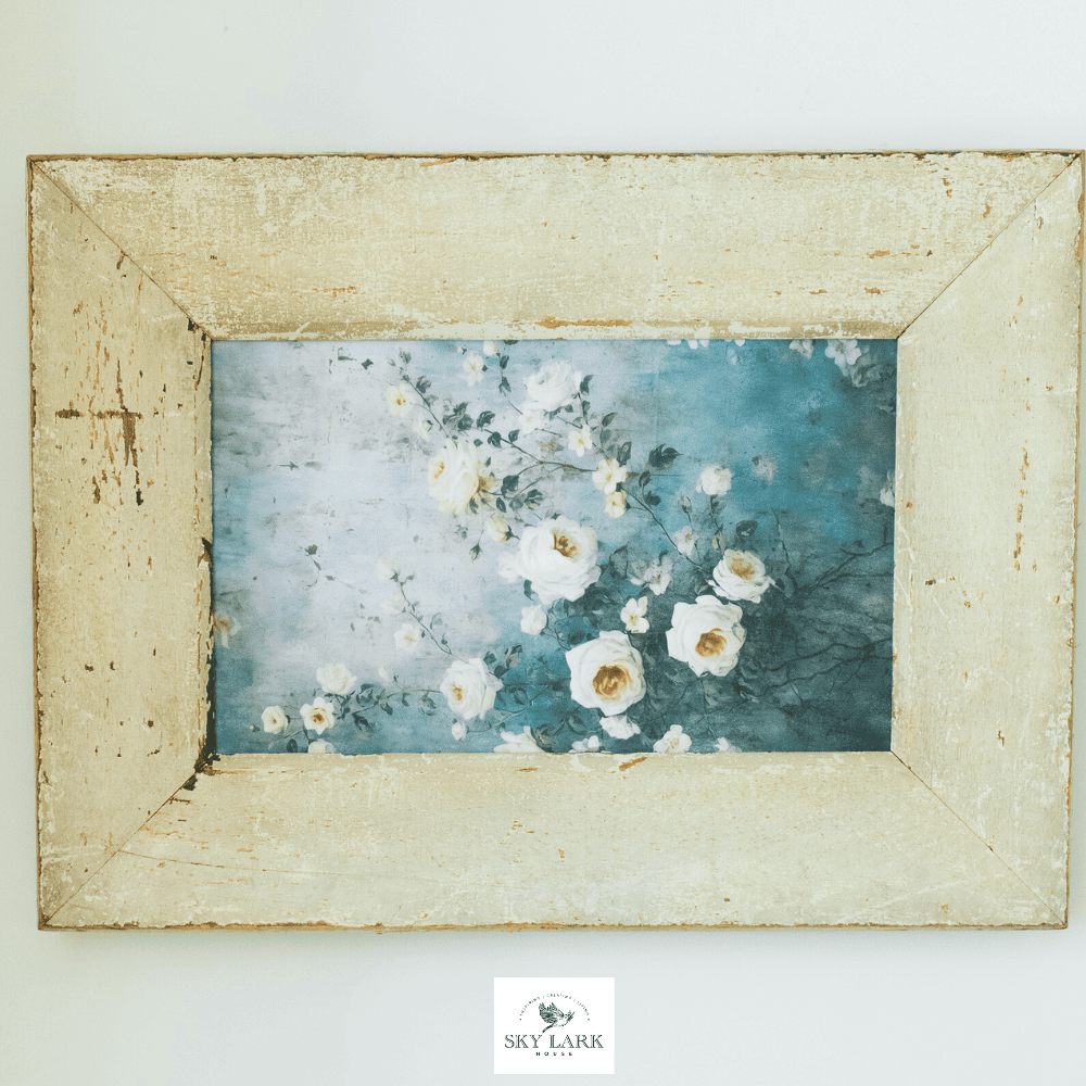 A rustic wooden frame surrounds a painting of white and pale pink roses with green leaves against a soft blue background. The artwork has a vintage, shabby-chic style.