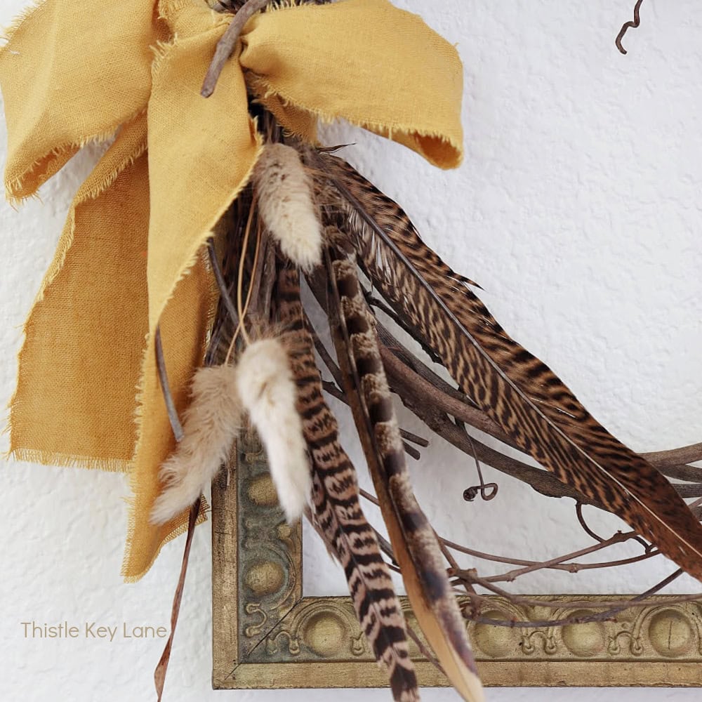 A rustic yellow fabric bow, dried grass, and striped brown feathers are attached to a wreath on a white textured wall, with part of an empty gold frame visible in the corner.