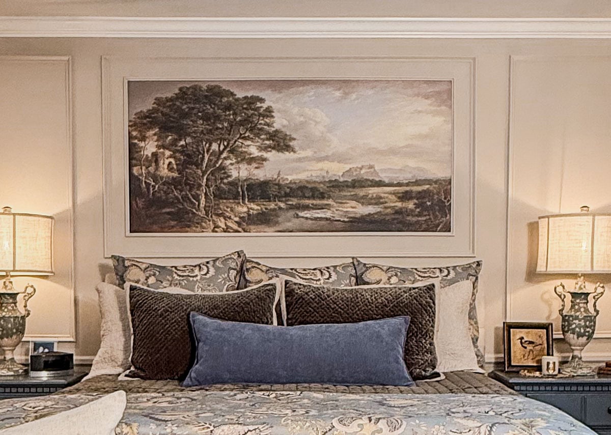A neatly made bed with patterned pillows and a blue cushion is centered below a large landscape painting—perfect inspiration for Master Bedroom Decorating Ideas. Two ornate lamps sit on nightstands with picture frames and small decor items.