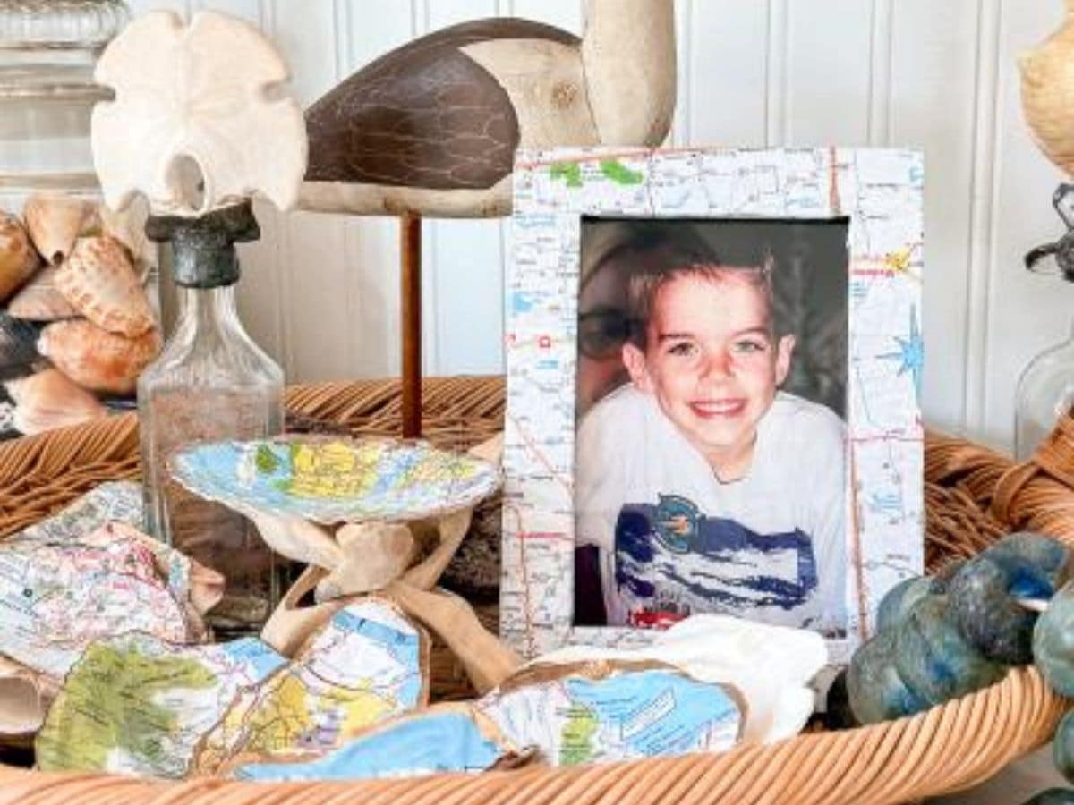 A wicker basket holds seashells, glass bottles, wooden bird figurines, and a framed photo of a smiling boy. The frame is decorated with pieces of a map, as are some scattered shells in the basket.