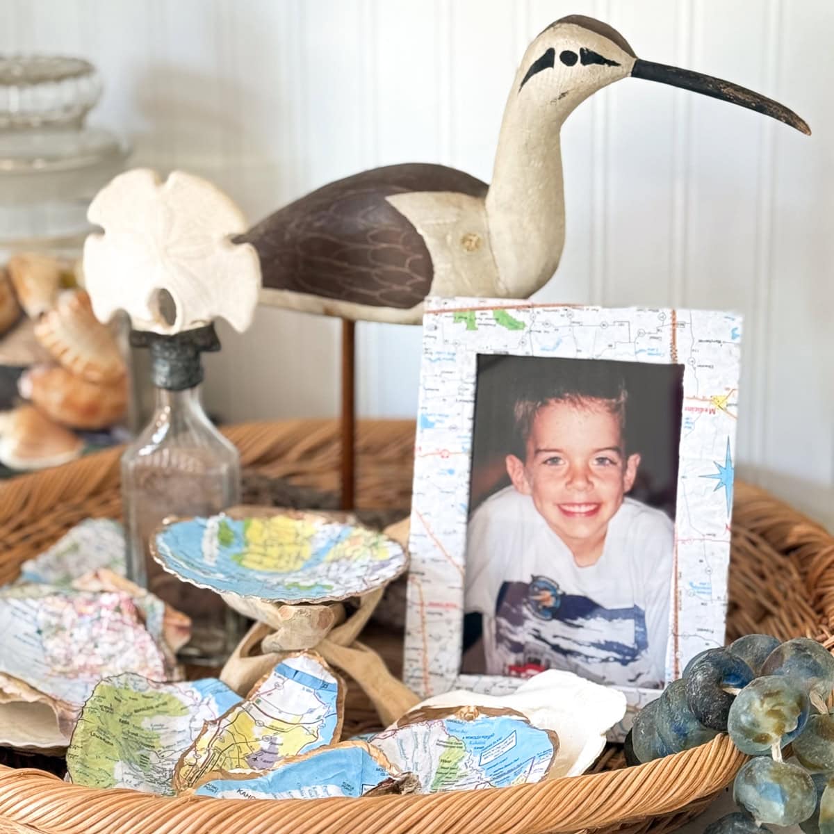 A wicker tray holds a wooden shorebird, a framed photo of a smiling boy, a sand dollar, a bottle with sand, seashells, and map-covered objects, with shells and grapes partially visible in the background.